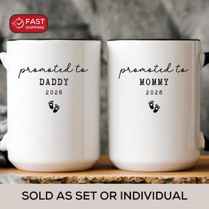 May include: Two white ceramic mugs with black handles and rims. One mug says "promoted to DADDY 2026" and the other says "promoted to MOMMY 2026" with baby footprints. Dishwasher and microwave safe.