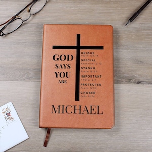 Puede incluir: Un diario de cuero marr&oacute;n con una cruz negra y el texto "GOD SAYS YOU ARE" con vers&iacute;culos b&iacute;blicos enumerados a continuaci&oacute;n. El nombre "MICHAEL" est&aacute; impreso en la parte inferior del diario.