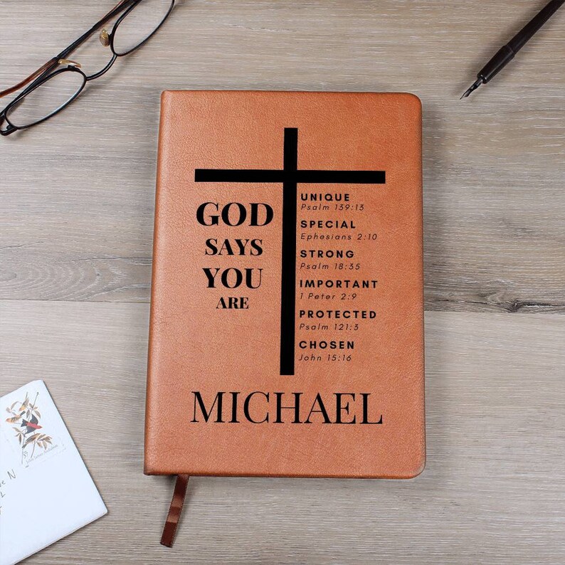 Puede incluir: Un diario de cuero marr&oacute;n con una cruz negra y el texto "GOD SAYS YOU ARE" con vers&iacute;culos b&iacute;blicos enumerados a continuaci&oacute;n. El nombre "MICHAEL" est&aacute; impreso en la parte inferior del diario.