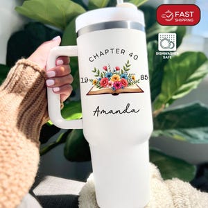 May include: White insulated tumbler with a handle, featuring a floral design and the text "Chapter 40 1985 Amanda." The tumbler has a white lid and handle. The design includes a book with colorful flowers.