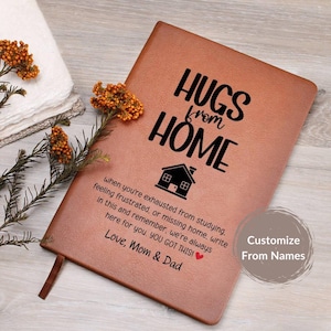 May include: A brown leather-bound journal with the words "HUGS from HOME" printed in black. The journal includes a house graphic and a message from parents. A ribbon bookmark is visible.