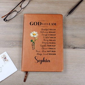 May include: A brown leather journal with the text "GODSAYS I AM" and a list of positive affirmations with corresponding Bible verses. A small daisy graphic is on the left side of the journal. The name "Sophia" is printed at the bottom of the journal.
