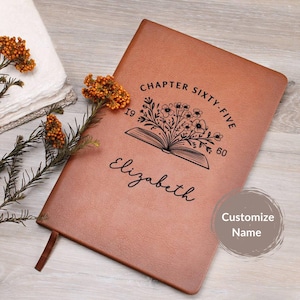 May include: A brown leather journal with the text "Chapter Sixty-Five 1960" and the name "Elizabeth" is displayed. The journal features an open book design with floral accents. A brown ribbon bookmark is visible.