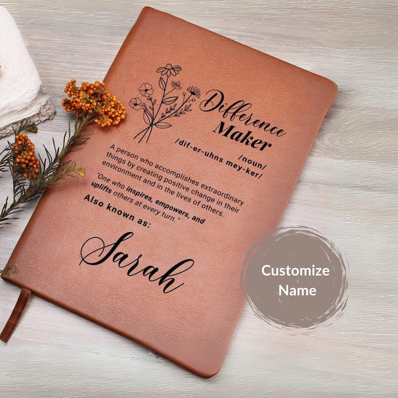 May include: A brown leather journal with the words "Difference Maker" defined as "a person who accomplishes extraordinary things by creating positive change in their environment and in the lives of others." The journal also has a floral design and the name "Sarah" written in cursive.