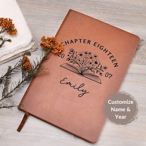 May include: A brown leather journal with the text "CHAPTER EIGHTEEN" and the name "Emily" printed on the cover. The journal features a floral design above an open book. A ribbon bookmark is visible.