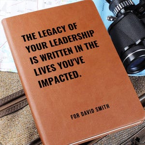 May include: A tan notebook with black text that reads: THE LEGACY OF YOUR LEADERSHIP IS WRITTEN IN THE LIVES YOU'VE IMPACTED. FOR DAVID SMITH. The notebook is positioned next to a pair of black binoculars and a map, resting on a textured surface.