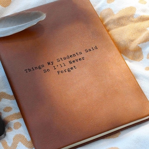 May include: A brown leather journal with the text "Things My Students Said So I'll Never Forget" printed on the cover. A white feather rests on the top left corner of the journal. The journal is closed and has a smooth texture.