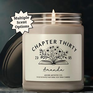May include: A lit, beige-colored candle in a glass jar with a silver lid. The label reads "CHAPTER THIRTY" with the name "Amanda" and the text "9 OZ SCENTED NATURAL SOY WAX CANDLE." A starburst graphic says "Multiple Scent Options."