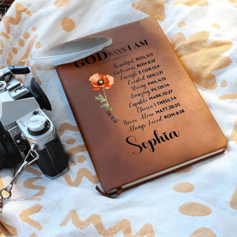 Puede incluir: Un diario de cuero marr&oacute;n con las palabras "Dios dice que soy" y una lista de afirmaciones positivas y vers&iacute;culos b&iacute;blicos. El diario est&aacute; decorado con una flor de amapola roja y el nombre "Sophia" est&aacute; escrito en la parte inferior.