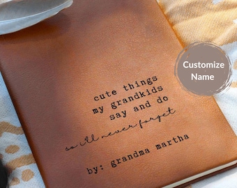 May include: A brown leather journal with the text "cute things my grandkids say and do so I'll never forget by: grandma martha." A feather rests on the top left corner. A customizable name button is in the top right corner.