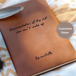 May include: A brown leather-bound notebook with the text "Documentation of the shit you can't make up" and "by michelle" in black script. A feather rests on the top left corner. A customizable name button is on the right.