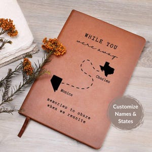 May include: A brown leather journal with the text "WHILE YOU were away" and "memories to share when we reunite." The journal features a map with two states and names. A brown ribbon bookmark is visible.