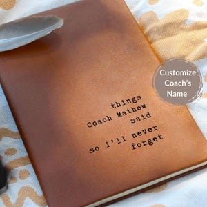 May include: A brown leather journal with the text "things Coach Mathew said so I'll never forget" printed on the cover. A feather rests on the top left corner. A circular graphic says "Customize Coach's Name".