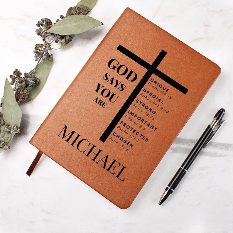 Puede incluir: Un diario de cuero marr&oacute;n con una cruz negra y el texto "GOD SAYS YOU ARE" con vers&iacute;culos b&iacute;blicos enumerados a continuaci&oacute;n. El nombre "MICHAEL" est&aacute; impreso en la parte inferior del diario.