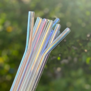 May include: A bundle of clear, iridescent glass straws. The straws are bent at the top and appear to have a rainbow sheen. The background is a soft, blurred green, suggesting an outdoor setting. These reusable straws are a sustainable alternative to plastic.