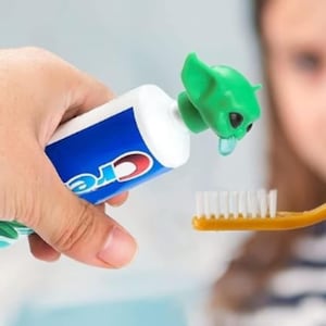 May include: A tube of Crest toothpaste with a green alien-shaped toothpaste dispenser. The dispenser is positioned over a yellow toothbrush, dispensing blue toothpaste. The tube is held by a hand.