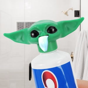 May include: A green character head with large ears and black eyes, attached to a tube of toothpaste. The toothpaste tube has a blue and white label with the brand name in red.