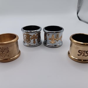May include: Four metal napkin rings. Two are gold-toned, one with an embossed design and the other with the initials "NMH". The other two are silver-toned with gold-colored cross and fleur-de-lis designs.