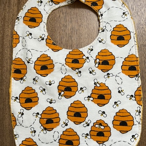 May include: A white baby bib with a yellow trim and a pattern of orange beehives and black bees. The bib has two yellow poppers at the neck.
