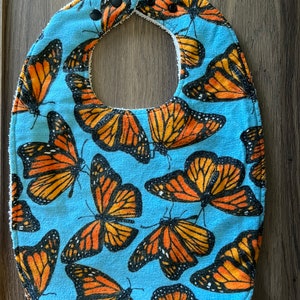 Puede incluir: Un babero azul para bebé con un patrón de mariposas monarca naranja y negras.
