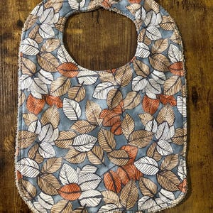 May include: A blue baby bib with a white and orange leaf pattern. The bib has a snap closure.