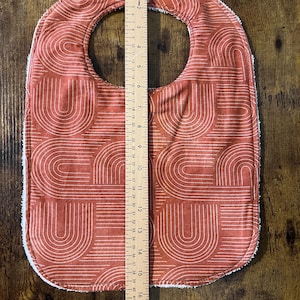 May include: A red and white baby bib with a geometric pattern of arches. The bib is made of soft, absorbent fabric and has a white trim around the edges.