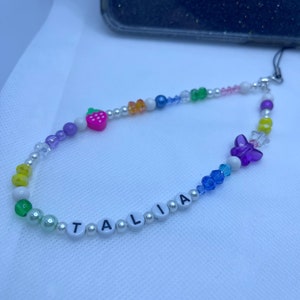 May include: A colourful beaded phone charm with the name "TALIA" spelled out in white letters. The charm features a variety of beads, including clear, purple, blue, green, yellow, and white. There is also a small strawberry charm and a butterfly charm.