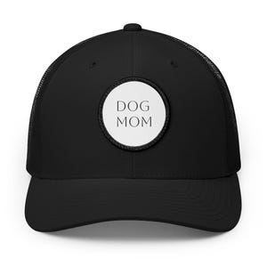 Könnte beinhalten: Eine schwarze Trucker-Cap mit einem runden weißen Aufnäher auf der Vorderseite. Der Aufnäher zeigt den Text DOG MOM in einer einfachen schwarzen serifenlosen Schrift. Die Kappe hat eine feste Vorderseite, einen gebogenen Schirm und ein Netz.