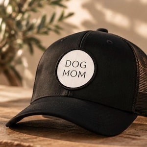 Könnte beinhalten: Eine schwarze Baseballkappe mit Netzrücken und einem runden weißen Aufnäher auf der Vorderseite. Der Aufnäher zeigt den Text DOG MOM in schwarzen Großbuchstaben. Die Kappe hat ein strukturiertes Vorderteil und einen gebogenen Schirm.