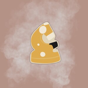 May include: A yellow ceramic chess piece shaped like a knight with white polka dots. The piece is standing on a light brown background.