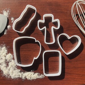 Pu&ograve; includere: Un set di sei tagliabiscotti in plastica bianca su una superficie di legno marrone. I tagliabiscotti hanno la forma di un cuore, una croce, un rettangolo, una tazza e due rettangoli di dimensioni diverse.