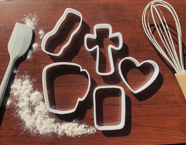 Pu&ograve; includere: Un set di sei tagliabiscotti in plastica bianca su una superficie di legno marrone. I tagliabiscotti hanno la forma di un cuore, una croce, un rettangolo, una tazza e due rettangoli di dimensioni diverse.