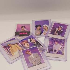 May include: A selection of K-pop idol photo cards in clear plastic sleeves. The cards show various members in different poses, with a purple and pink backdrop. Some cards have text in a foreign language.