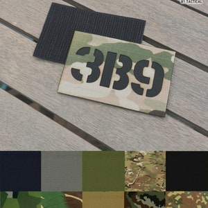 May include: A rectangular patch displaying the number "389" in black stencil font on a camouflage pattern. A separate black hook-and-loop fastener patch is above it. A selection of fabric swatches in various colours is also shown.