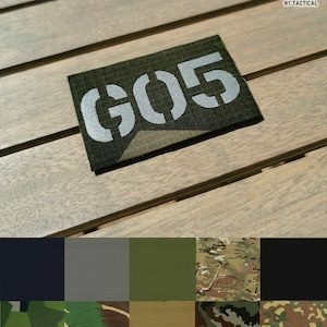 Könnte beinhalten: Ein rechteckiger, olivgrüner Aufnäher mit dem weißen Schablonentext "G05". Der Aufnäher wird auf einer Holzoberfläche angezeigt. Das Bild zeigt auch eine Farbpalette von Stoffmustern in verschiedenen Grüntönen, Schwarz und Tarnmustern. Das Logo "N1 TACTICAL" befindet sich in der oberen rechten Ecke.