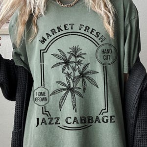 May include: Olive green t-shirt with a black graphic design. The design features a stylized plant illustration within a rectangular frame, with the words "MARKET FRESH", "HOME GROWN", "HAND CUT", and "JAZZ CABBAGE".