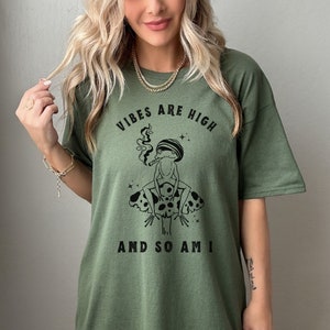 May include: Green t-shirt with a black and white graphic of a frog smoking on a mushroom. The text "Vibes are high and so am I" is printed on the shirt.