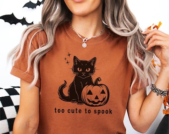 May include: A brown t-shirt with a black cat and pumpkin graphic. The text "too cute to spook" is printed below the graphic.