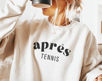 May include: A woman wearing a cream-colored sweatshirt with the text "après tennis" in black lettering. She is sitting on a couch with her legs crossed and holding a black mug.