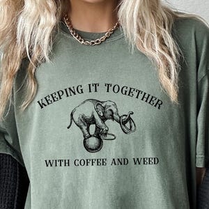 May include: Olive green t-shirt with the text "KEEPING IT TOGETHER" above an illustration of an elephant holding a coffee cup and a cannabis plant. Below the illustration is the text "WITH COFFEE AND WEED".