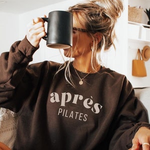 May include: A woman wearing a brown sweatshirt with the text "apr&egrave;s PILATES" in white. She is sitting on a couch with her legs crossed and holding a black mug.
