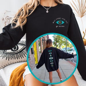 May include: Black sweatshirt with the text "May every evil eye go blind" and a blue eye graphic on the front. The back of the sweatshirt features a vertical row of blue eyes.