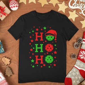 May include: Black t-shirt with a Christmas-themed design featuring the words "HO HO HO" and three red and green pickleball paddles with a Santa hat on top of the first paddle.