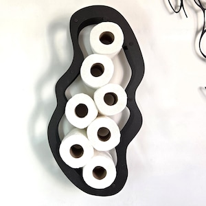 May include: A black, organically shaped toilet paper holder filled with seven rolls of white toilet paper. The holder is mounted on a white wall, with a black abstract art piece in the background.