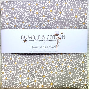 May include: A gray flour sack towel with a white daisy pattern and yellow centers. The towel has a white label with the text "BUMBLE & COTTON nature & whimsy homewares Flour Sack Towel 20 x 30 | 100% cotton | handmade in oregon".