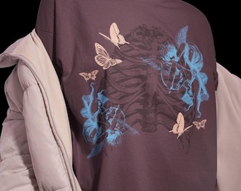 May include: A dark gray t-shirt featuring a graphic design of a rib cage, butterflies, and cherubs in shades of blue and beige. The shirt is worn with a light gray puffer jacket.