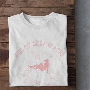 May include: White t-shirt featuring a pink graphic design of a woman sitting on a planet with a rocket ship and stars. The text "out of this world" is written in a pink cursive font.