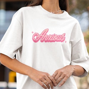 May include: White t-shirt with the word "Anxious" in pink, retro-style lettering. The word is slightly curved and has a shadow effect, with small starbursts on either side. The shirt has short sleeves and a relaxed fit.