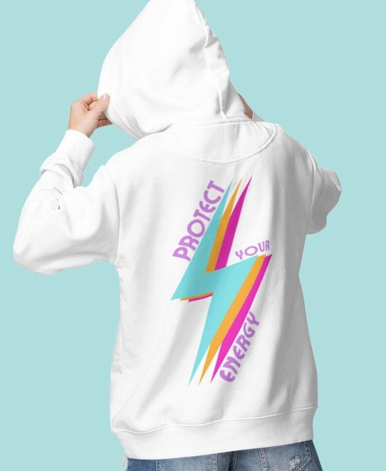 K&ouml;nnte beinhalten: Wei&szlig;er Hoodie mit einem Blitz-Motiv und dem Text "Sch&uuml;tze deine Energie" in Lila und Orange.