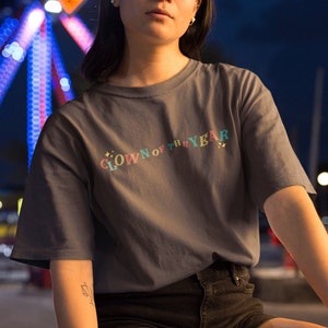 May include: A dark gray t-shirt with the text "CLOWN OF THE YEAR" in rainbow colors. The text is in a curved arc with stars on either side.